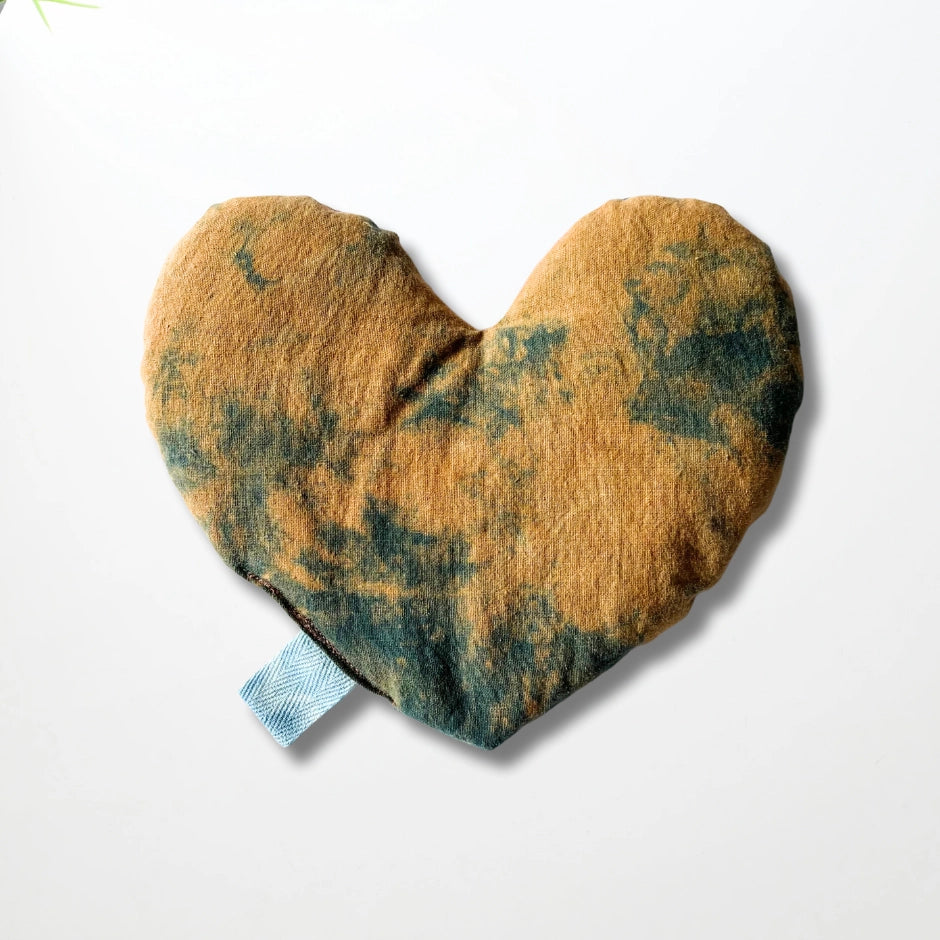 The Faire Heart Eye Pillow is a heart-shaped cotton pillow with a brown and dark green tie-dye pattern and a small blue tab—ideal for gentle anxiety relief. Shown on a plain white background.