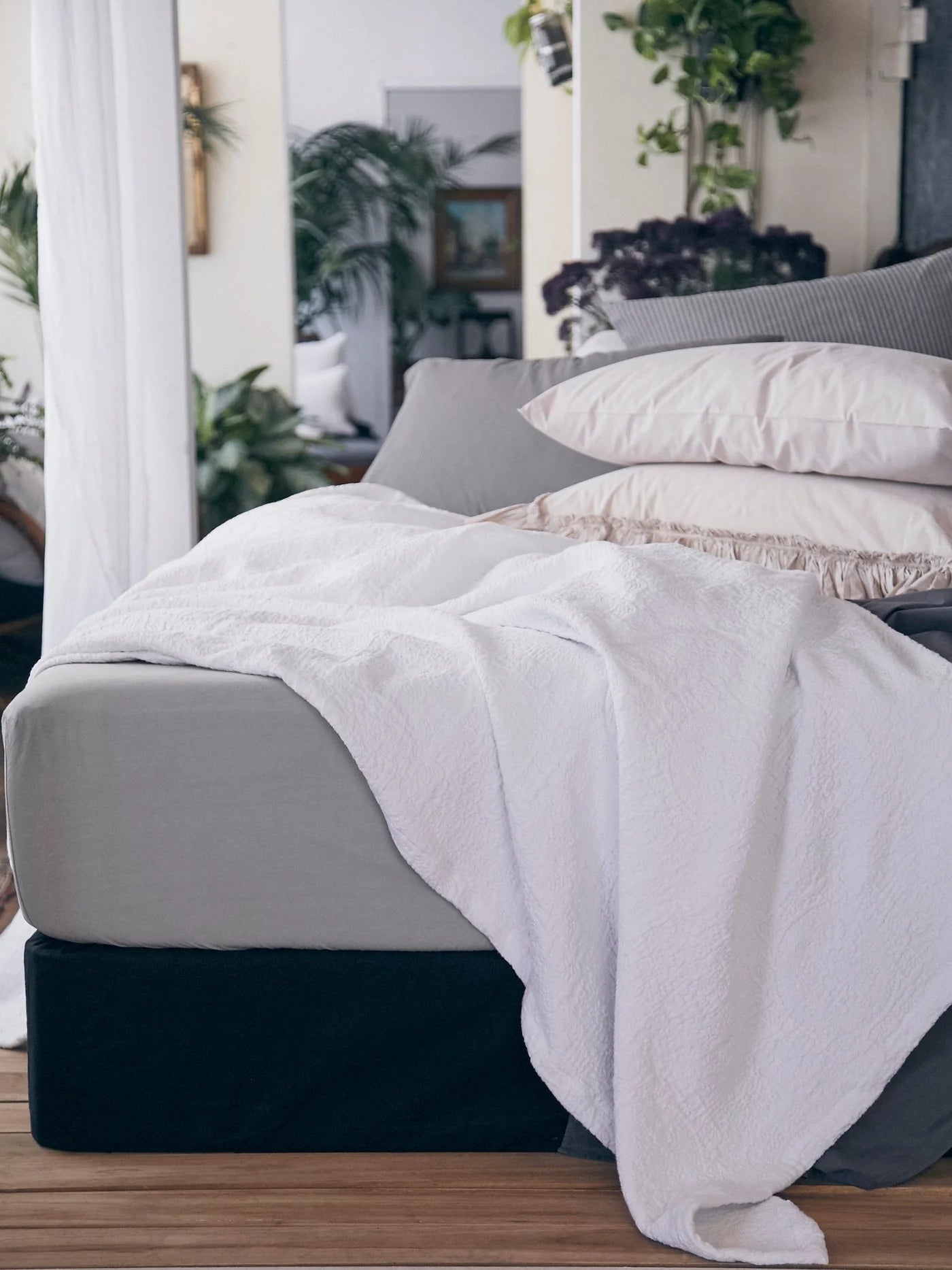 A neatly made bed features Matteo's Antik Matelasse Coverlet in white, paired with light gray sheets and white pillows. Green plants accent the room, creating a cozy, natural atmosphere.