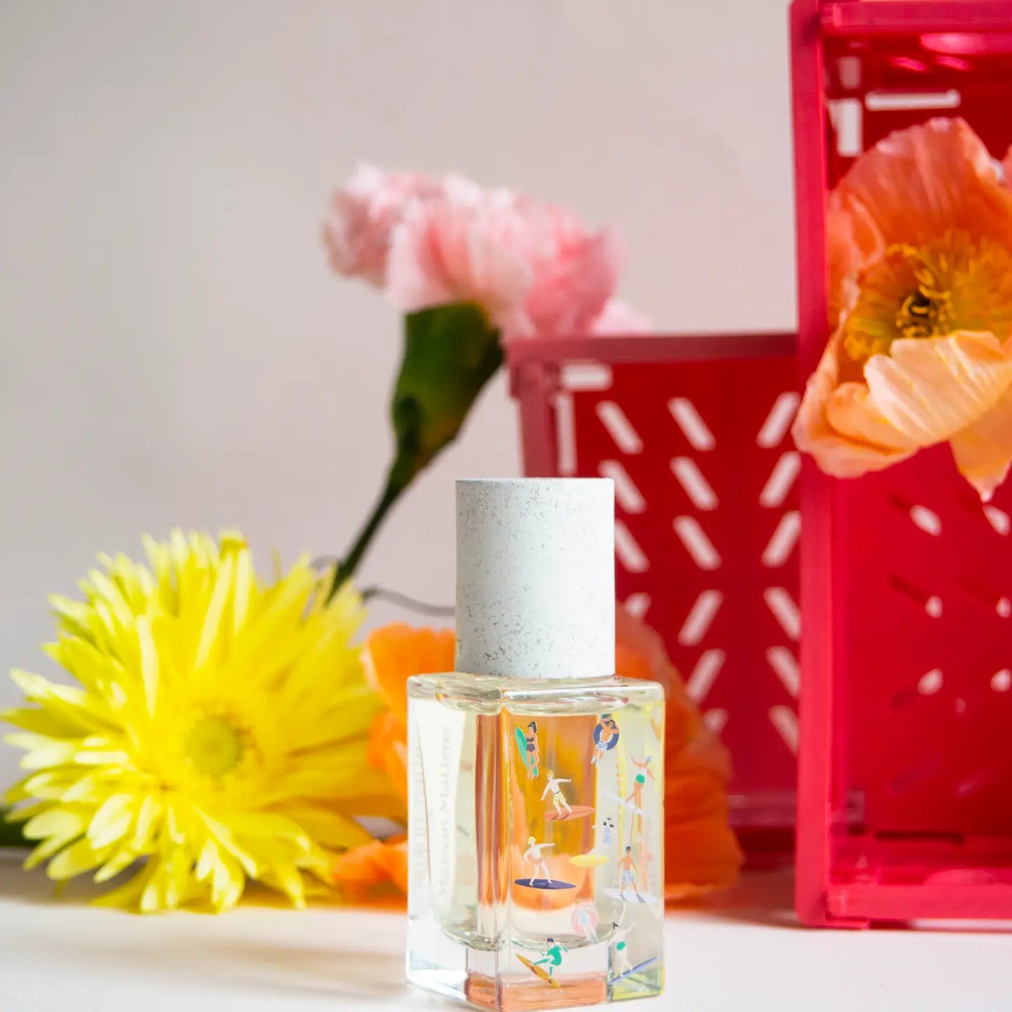 The Maison Matine Perfume by Faire, featuring a white cap and delicate designs, is displayed before pink, yellow, and orange flowers that evoke soft vanilla notes. A red crate appears in the background on a light surface.