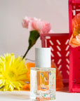The Maison Matine Perfume by Faire, featuring a white cap and delicate designs, is displayed before pink, yellow, and orange flowers that evoke soft vanilla notes. A red crate appears in the background on a light surface.