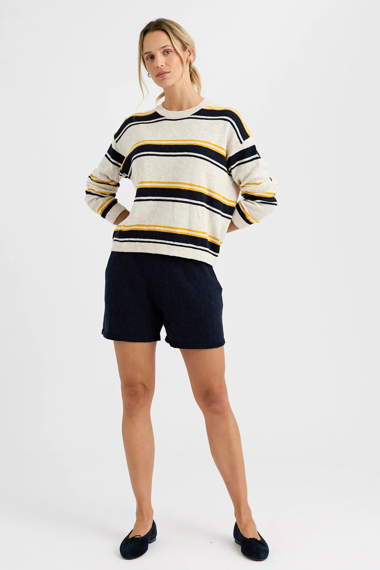A woman stands with her hands in the pockets of dark navy shorts, wearing the Susannah Pullover by One Grey Day—a cream sweater with black and mustard stripes—paired with black flats, set against a plain white background.