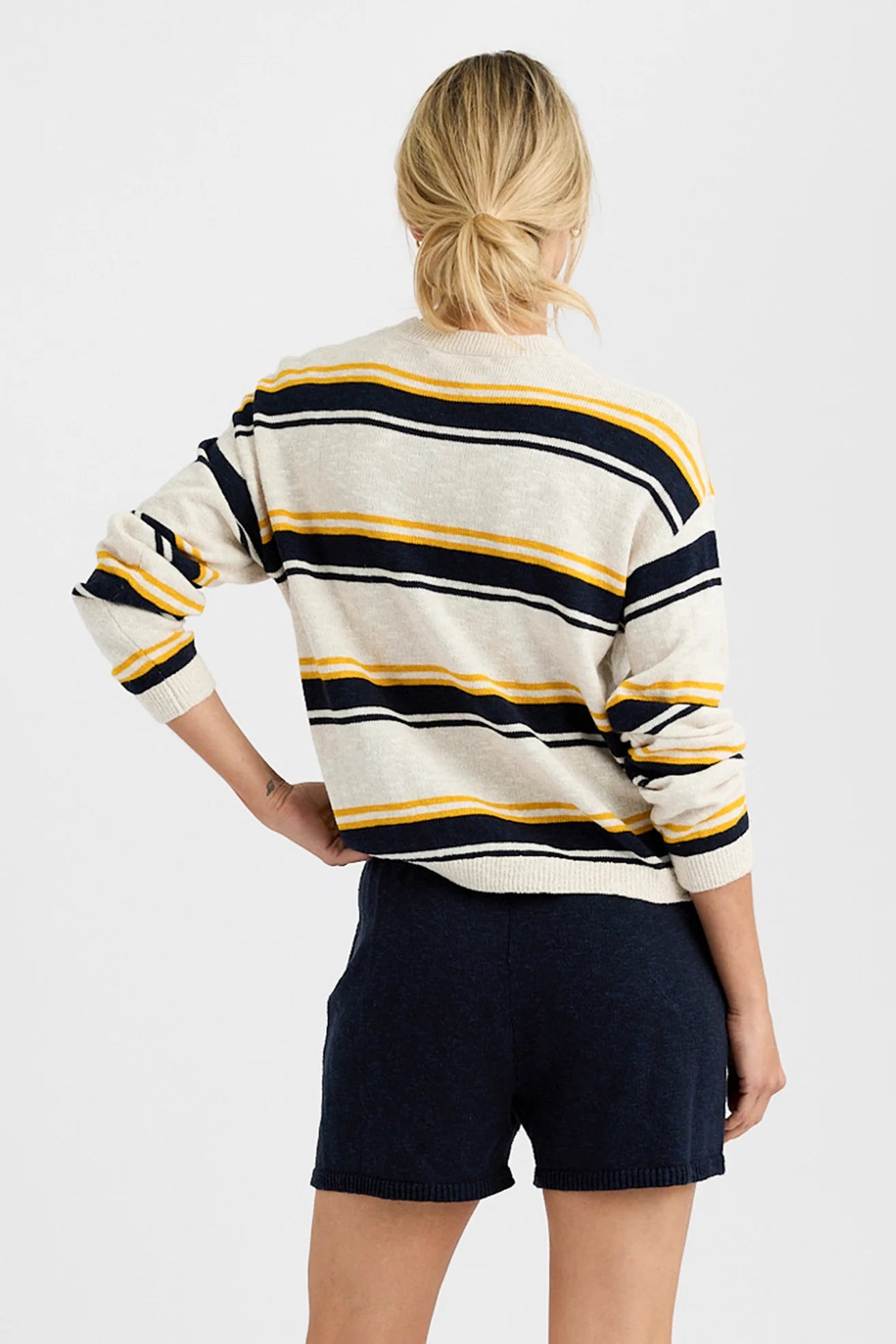 Seen from behind against a plain light background, a person with blonde hair in a low bun wears the One Grey Day Susannah Pullover—a striped sweater—paired with dark navy shorts.