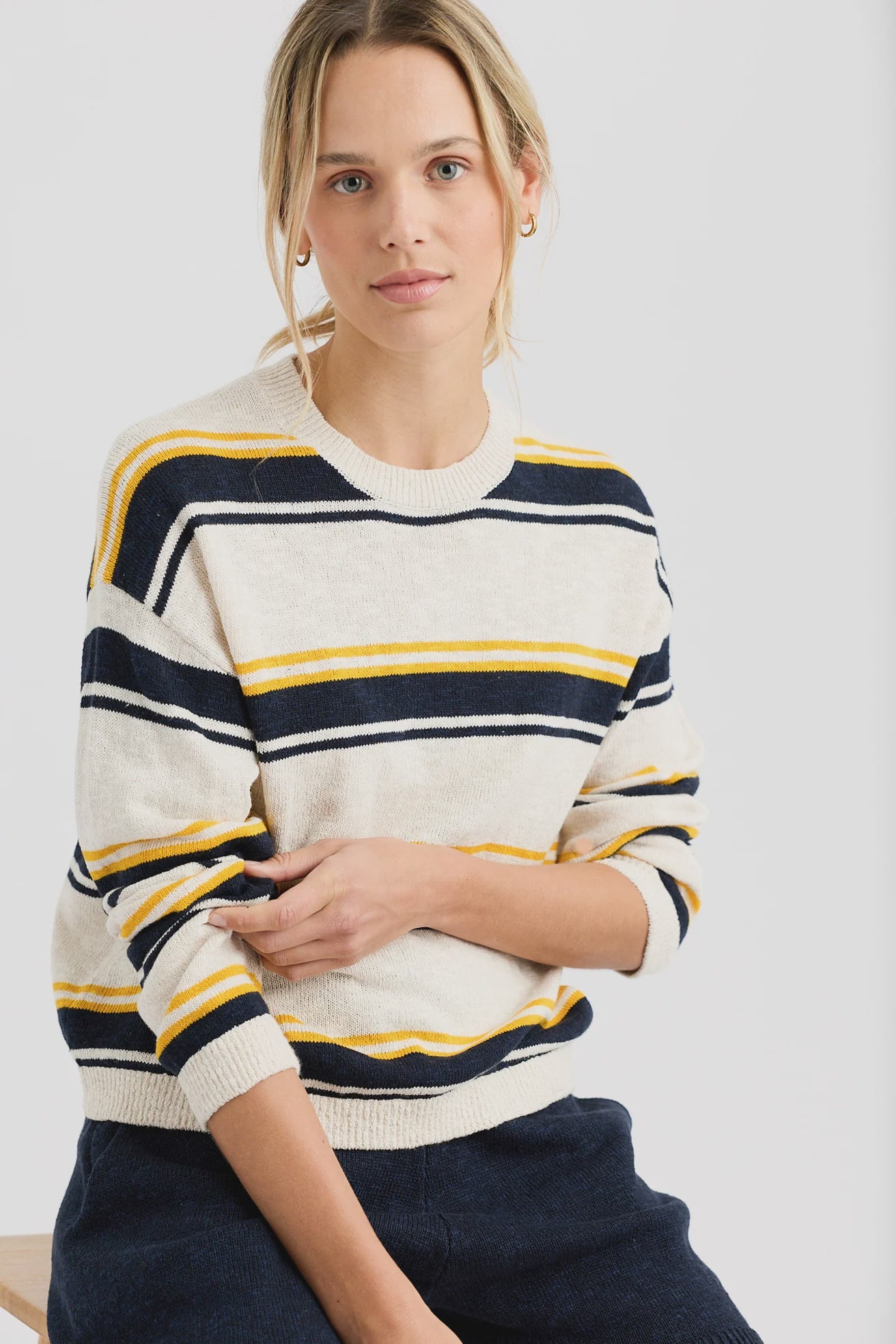 A woman with blonde hair wears the Susannah Pullover by One Grey Day, a striped sweater in cream with navy and yellow stripes, as she sits and looks at the camera against a plain white background.
