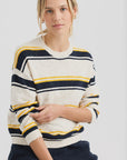 A woman with blonde hair wears the Susannah Pullover by One Grey Day, a striped sweater in cream with navy and yellow stripes, as she sits and looks at the camera against a plain white background.