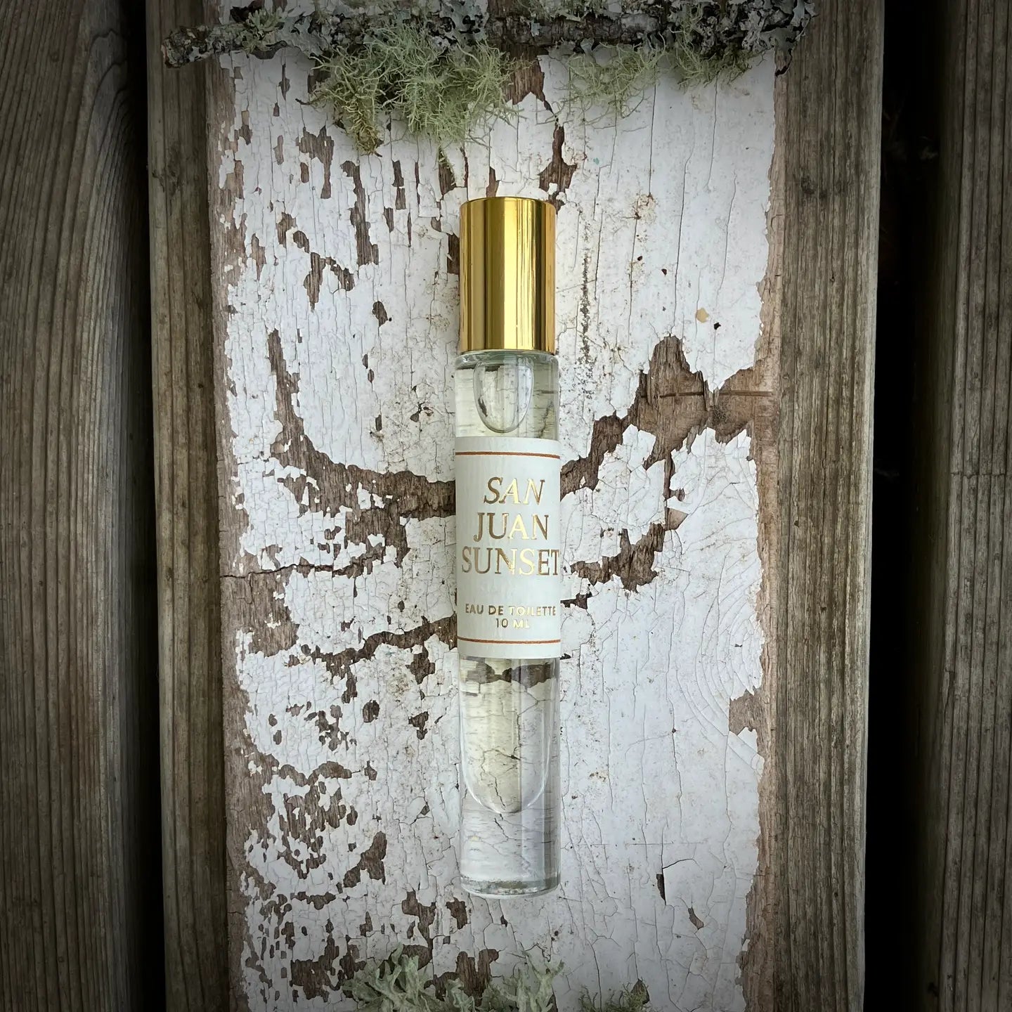 A glass "San Juan Roll On" perfume by Faire with a gold cap rests on weathered white paint and mossy wood, evoking the citrus-kissed breeze of the San Juan Islands.
