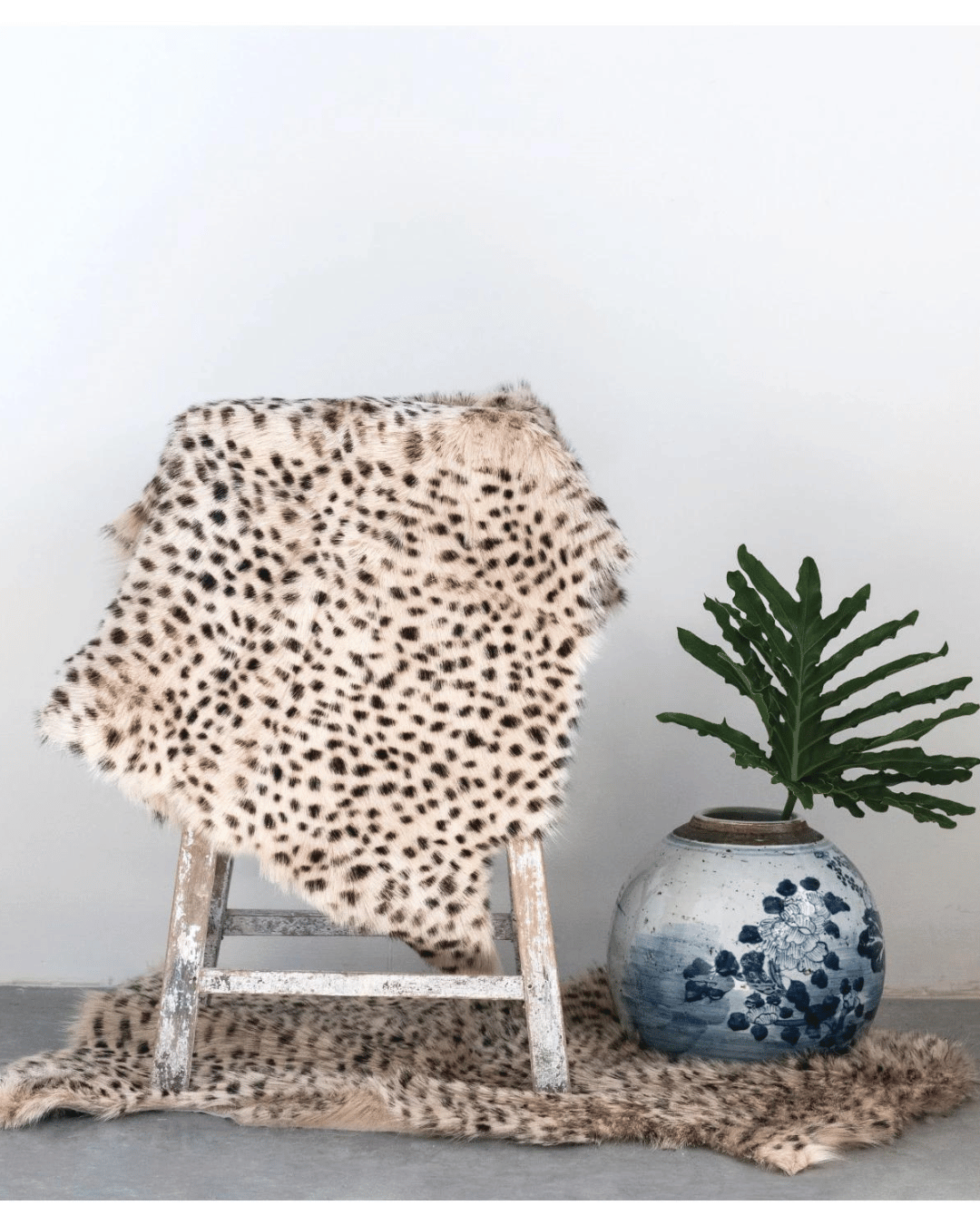 A small wooden stepladder draped with a luxurious Creative Co-op Goat Fur Rug Leopard Print sits beside a large ceramic pot with blue floral designs. The pot contains a lush, green plant, and another silky rug lies on the floor beneath the ladder.