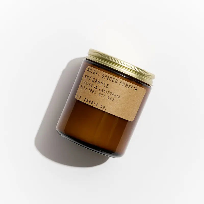 A brown glass jar PFC Candle by Faire with a gold lid casts a shadow. Its kraft label reads "Spiced Pumpkin Soy Candle, 100% soy wax, hand-poured, made in California" on a white background.