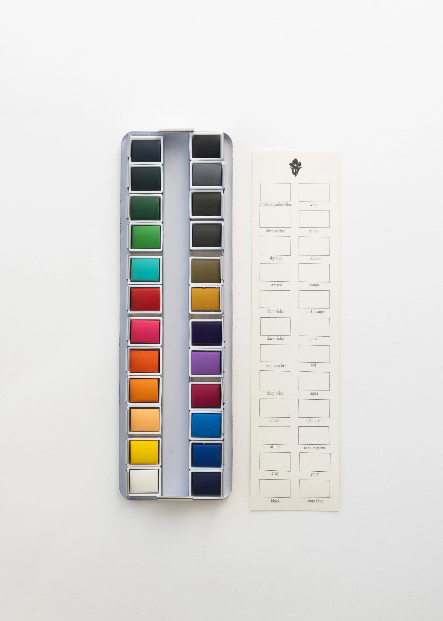 The Faire Watercolor Paint Palette features 18 paint pans arranged in a metal case, shown beside a cream label sheet with black printed color names on a white background.