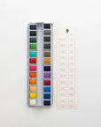 The Faire Watercolor Paint Palette features 18 paint pans arranged in a metal case, shown beside a cream label sheet with black printed color names on a white background.