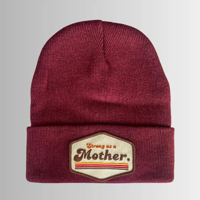 The Faire Patch Beanie Strong as a Mother is a maroon plush beanie with an adjustable cuff and a beige patch on the front that reads "Strong as a Mother" in red and black, accented by red stripes.