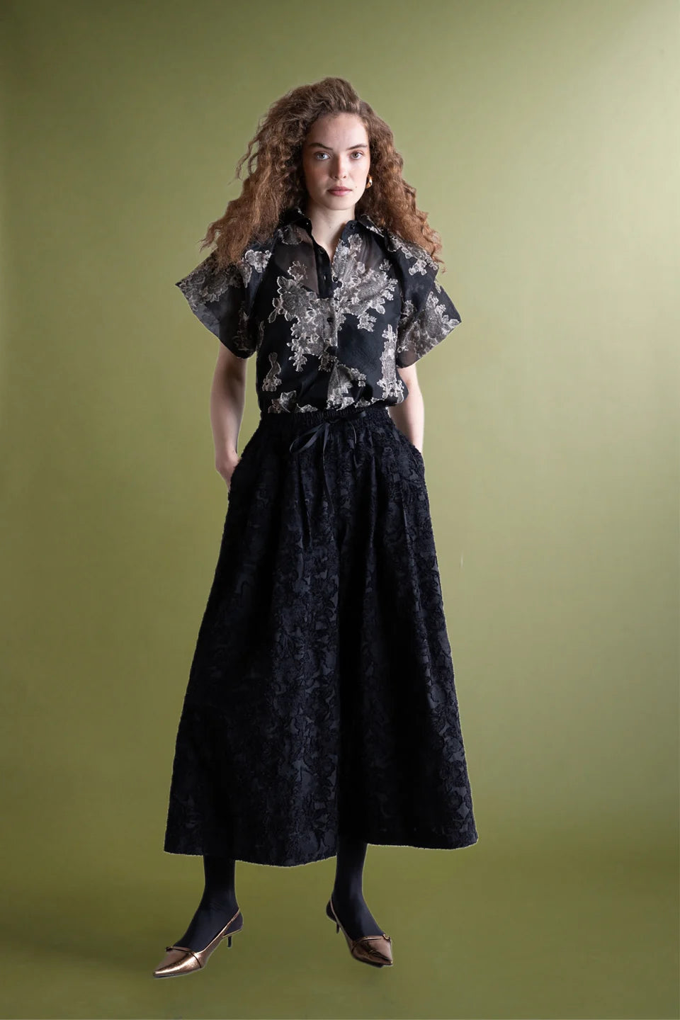 A woman with long curly hair stands against a plain olive green background, wearing a black short-sleeve floral blouse, Palazzo Wide Leg Trouser by Psophia, black tights, and pointed metallic heels.