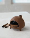 The Creative Co-op Footed Incense Chimney is a small, terra-cotta colored ceramic holder with a rounded oven shape on a white surface. It emits fragrant smoke from burning incense and features burned sticks beside it, all enhanced by its charming reactive glaze finish.