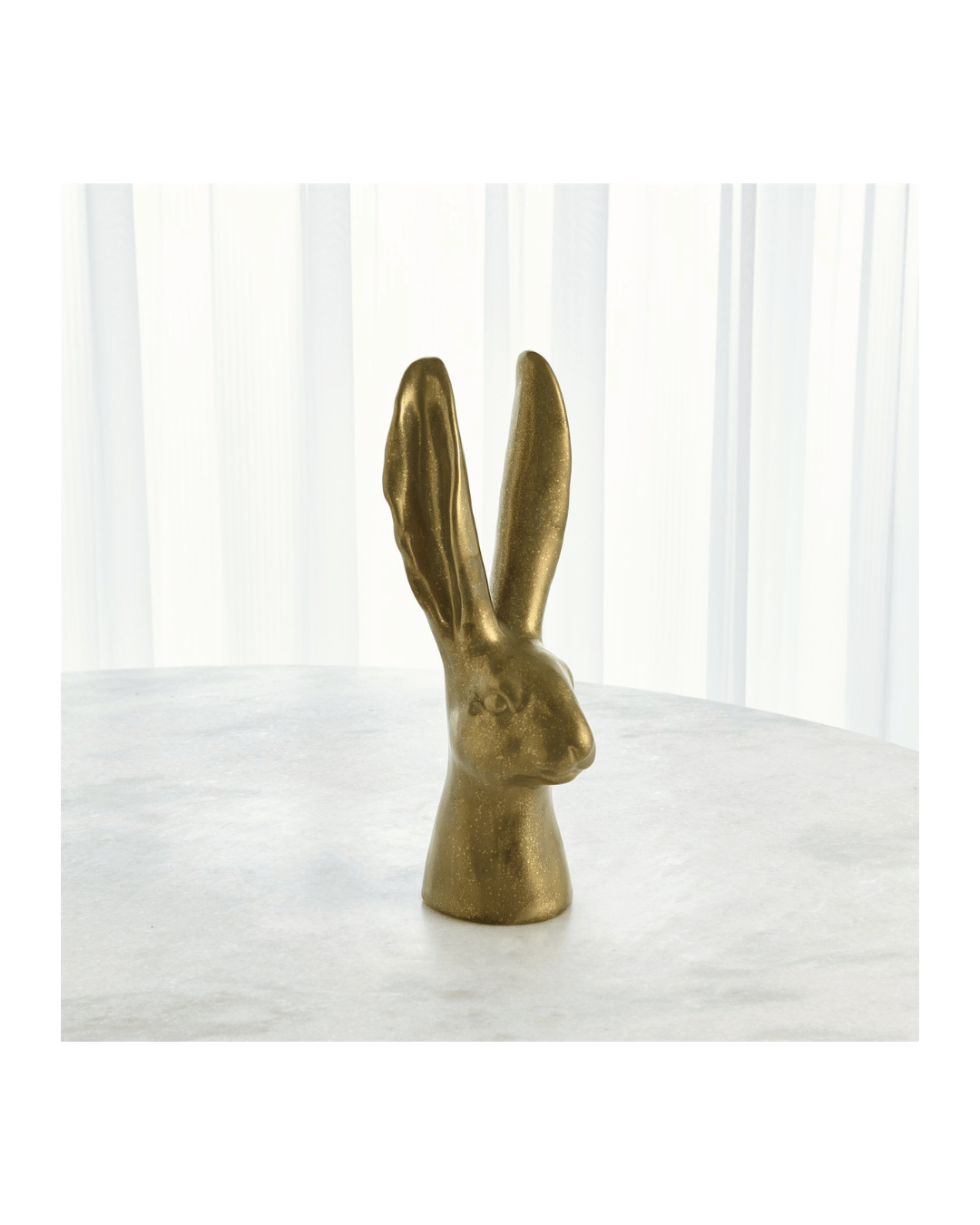 The Global Views Rabbit-Matte Gold Medium sculpture, featuring a ceramic rabbit head with elongated ears, rests elegantly on a light marble surface. The elegant scene is complemented by softly diffused vertical white curtains in the background.