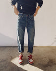 A person wearing a black long-sleeve shirt, The Loose Crop Selvedge jeans by Hey Gang with rolled cuffs, and red and white sneakers is standing on a concrete floor in natural lighting. The person's face is not visible.