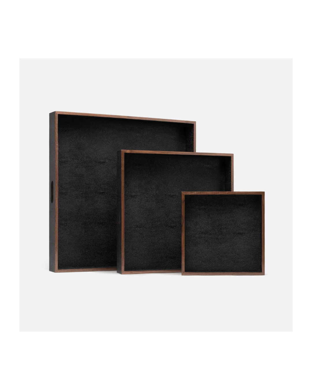 The Amina Trays by Made Goods feature a set of three rectangular trays, each with a dark interior and a natural wood finish on the edges. They are artfully stacked in a nested arrangement against a white background, offering an elegant touch to any setting.