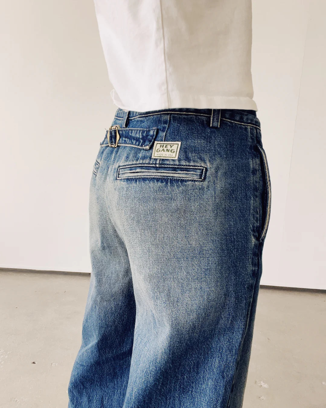 A person dressed in Hey Gang's The Buckle Back Trouser and a white shirt stands in a minimalistic indoor setting. Made in Los Angeles, the slightly faded cotton denim trousers feature a visible label on the back pocket, adding a vintage look against the plain white wall.