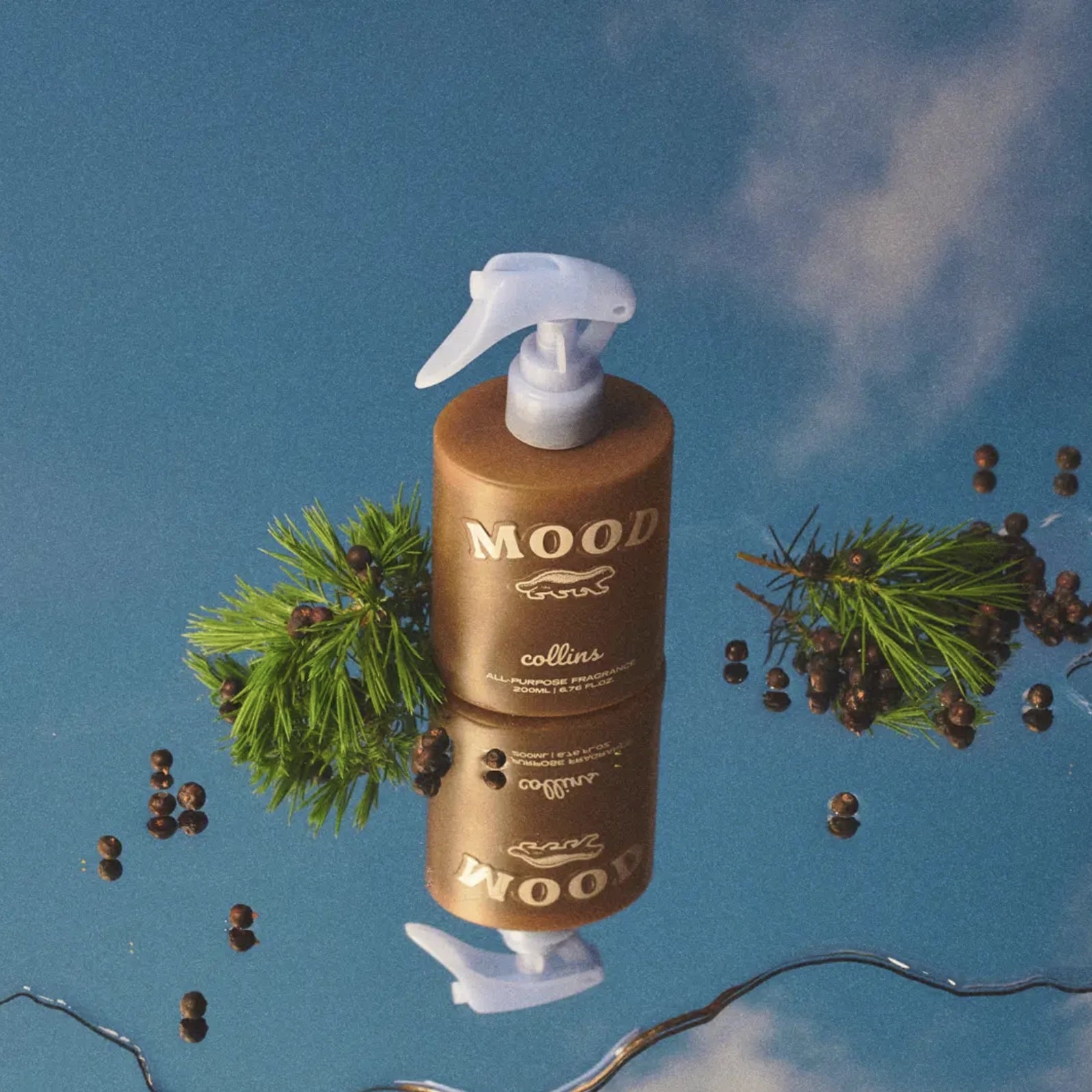 A brown "Scottish Juniper Room Spray" by Faire rests on a reflective surface with pine branches and berries, set against a sky-like backdrop—an inviting ethanol-free fragrance mist for any room.