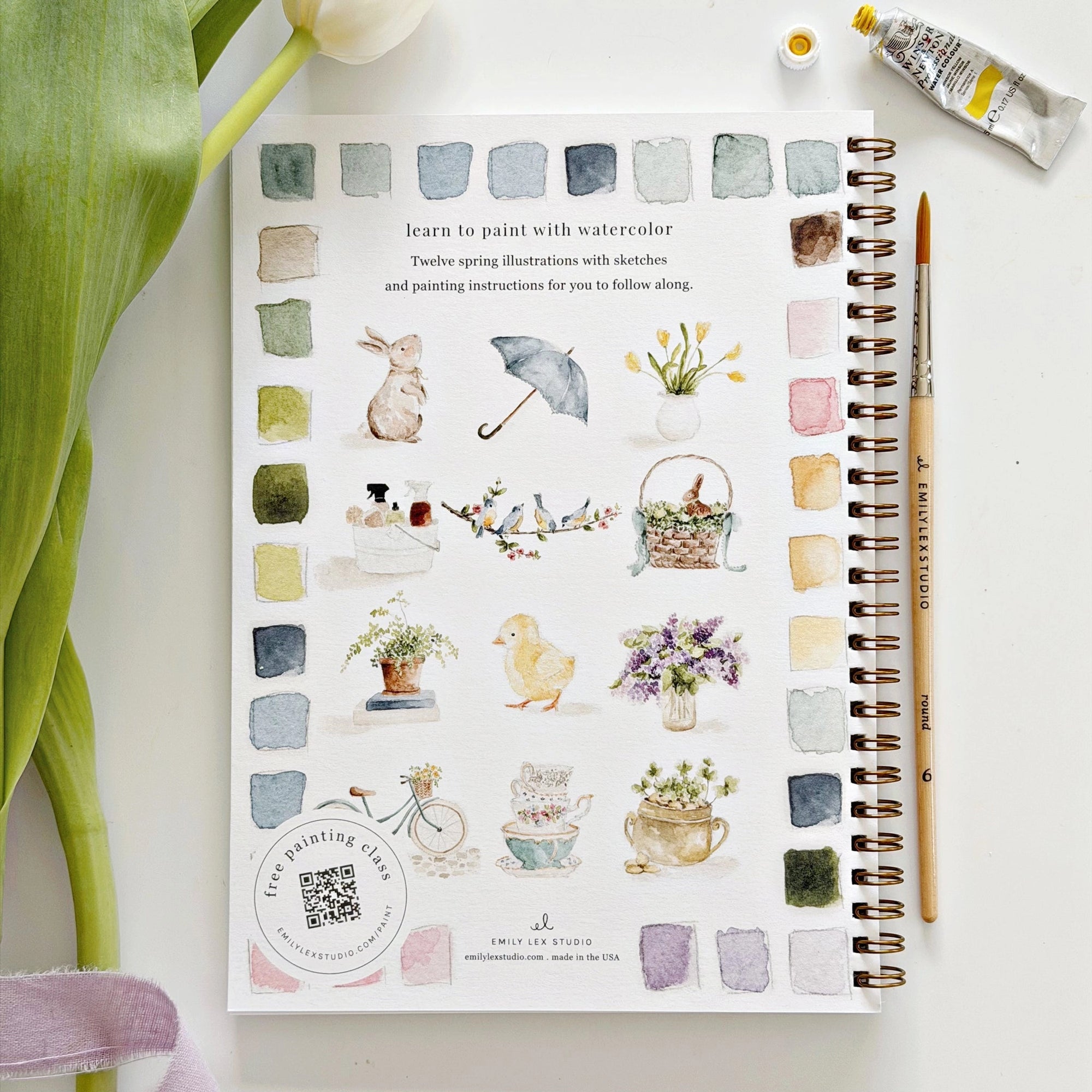 The Faire Spring watercolor workbook rests on a white surface, displaying spring-themed illustrations like animals, flowers, and teacups with color swatches. Ideal for beginner art activities; a paintbrush, paint tube, and green tulip leaf are nearby.