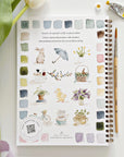 The Faire Spring watercolor workbook rests on a white surface, displaying spring-themed illustrations like animals, flowers, and teacups with color swatches. Ideal for beginner art activities; a paintbrush, paint tube, and green tulip leaf are nearby.
