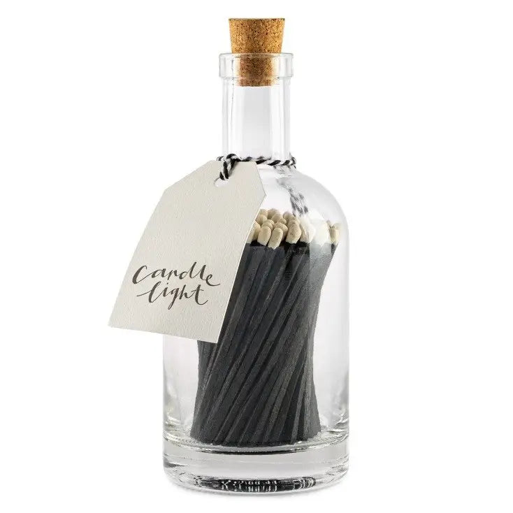 Faire's Glass Bottle Matches feature black matches in a clear glass bottle with a cork stopper, promoting sustainable home living. A string-attached white tag reads "Candle Light," highlighting its dedication to plastic-free packaging.