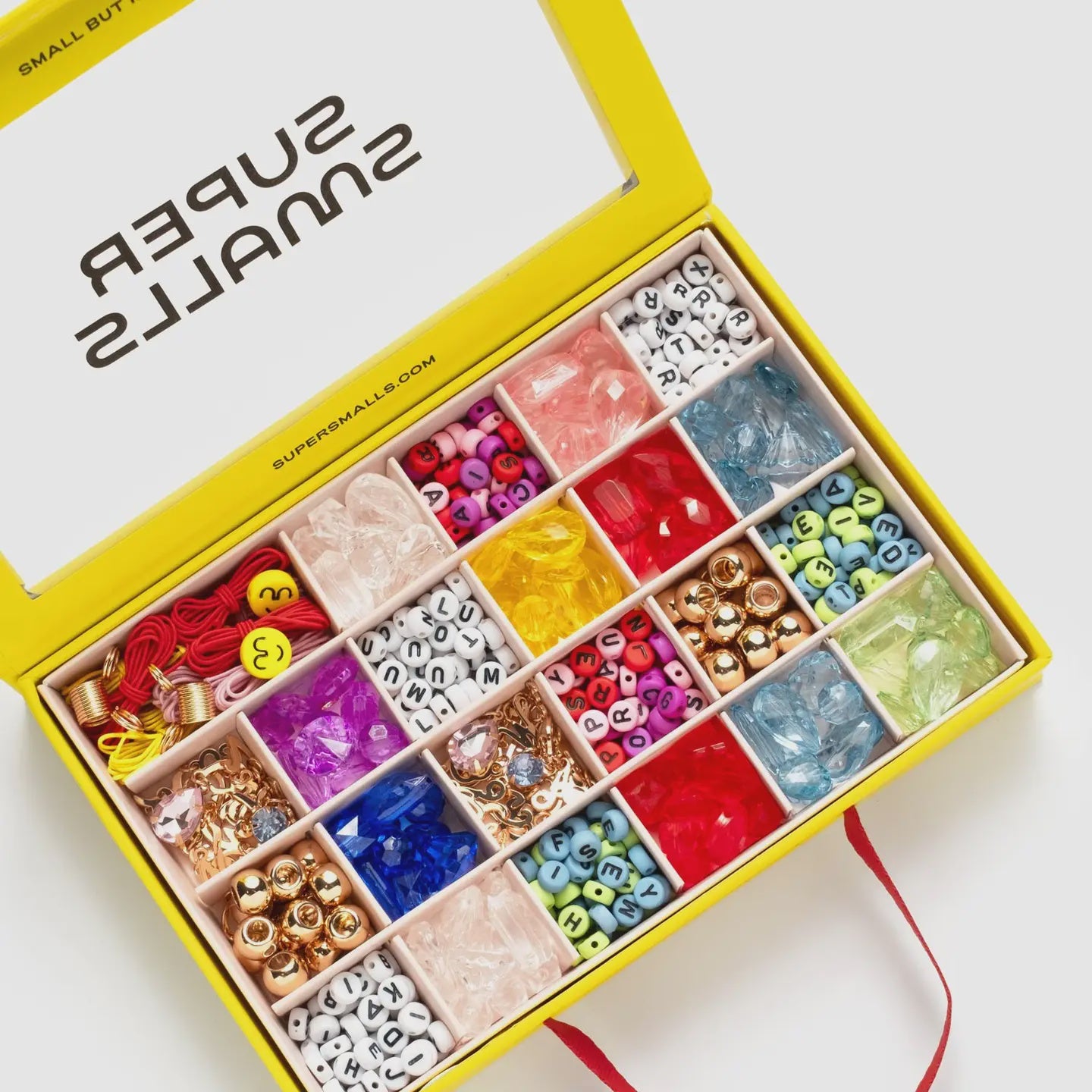 A yellow box labeled "Make It Alphabet DIY Bead Kit" by Faire contains colorful compartments of beads, letters, and baubles in various shapes and sizes. The lid features a red ribbon for closure.