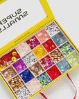 A yellow box labeled "Make It Alphabet DIY Bead Kit" by Faire contains colorful compartments of beads, letters, and baubles in various shapes and sizes. The lid features a red ribbon for closure.