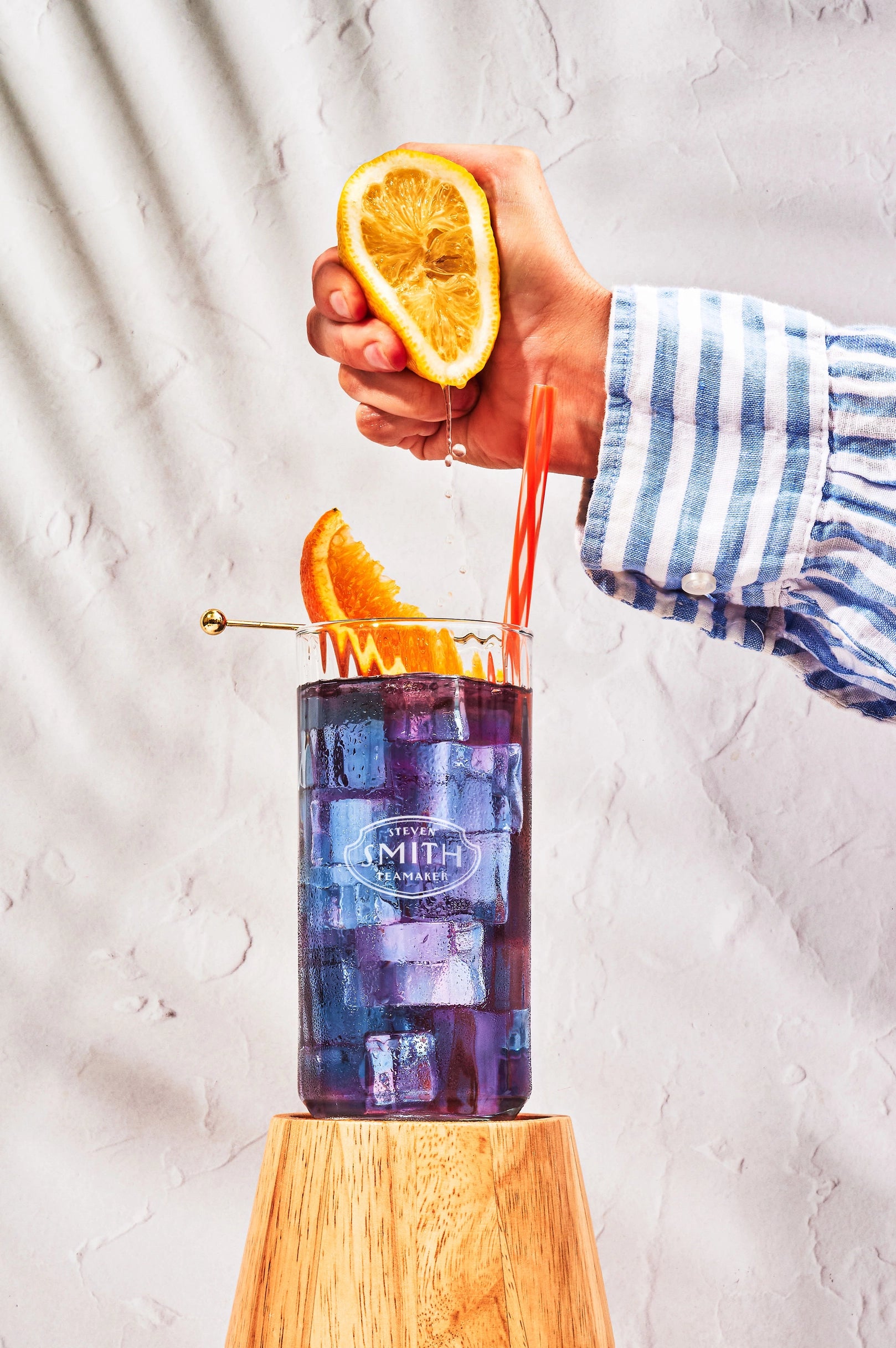 A hand in a blue and white striped shirt squeezes a lemon half over a tall glass of Faire Blackberry Jasmine Iced Tea with ice and orange slices, set on a wooden pedestal against a textured white background.