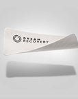 A white Dream Strips sleep mask by Faire, crafted from bamboo silk with "Dream Recovery" and a circular design, is set against a light gray backdrop, casting a soft shadow. Slightly folded on one side, it's an elegant touch for better sleep habits.