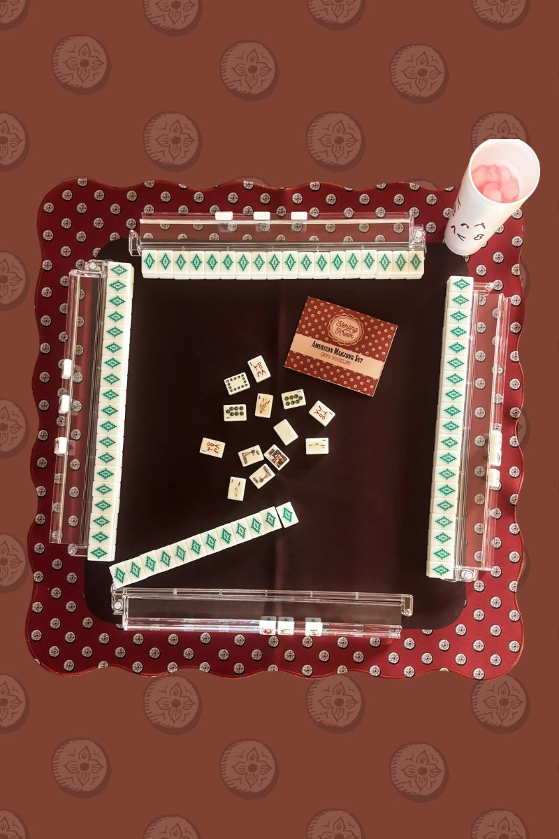 A Faire Wild West Mahjong Mat ONLY in red pattern, shown from above with tiles, racks, a smiling face cup with pink contents, and a red booklet.