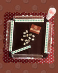 A Faire Wild West Mahjong Mat ONLY in red pattern, shown from above with tiles, racks, a smiling face cup with pink contents, and a red booklet.