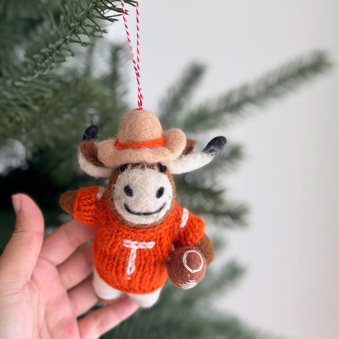 A hand holds the Faire Felted Ornament—a Christmas tree decoration shaped like a smiling longhorn cow in a cowboy hat and orange "T" sweater, holding a football, adding festive charm to your holiday décor.