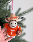 A hand holds the Faire Felted Ornament—a Christmas tree decoration shaped like a smiling longhorn cow in a cowboy hat and orange "T" sweater, holding a football, adding festive charm to your holiday décor.