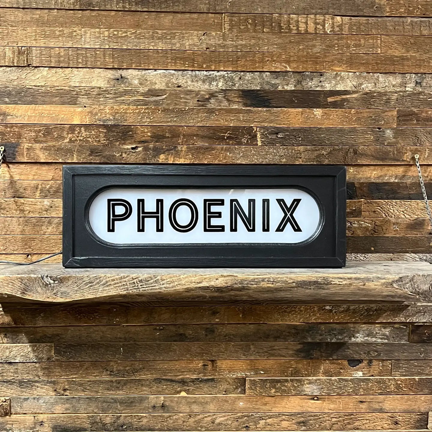 The Faire Lighted Sign features bold black “PHOENIX” lettering on a white background, displayed on a rustic wooden shelf against plank walls, capturing the vintage charm of a classic marquee.