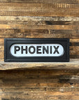 The Faire Lighted Sign features bold black “PHOENIX” lettering on a white background, displayed on a rustic wooden shelf against plank walls, capturing the vintage charm of a classic marquee.