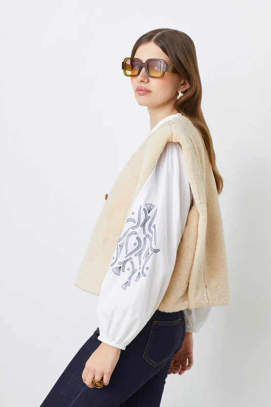 A woman with long brown hair wears large square sunglasses, the SUNCOO Diego Vest in cream recycled polyester, an embroidered-sleeve white blouse, and dark blue jeans, standing against a plain white background.