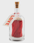 The Glass Bottle Matches by Faire feature a clear glass container with red-tipped matches, promoting sustainable living with plastic-free packaging. A cork stopper secures the bottle, and a string-tied tag reads "LOVE" at its neck.