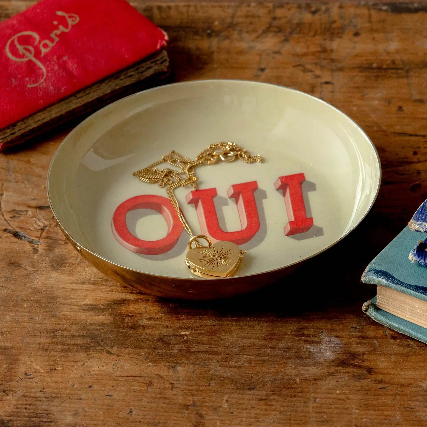 Gold heart locket necklace in a round enamel tray by Bungalow Furniture, displayed on a wooden table with books nearby.