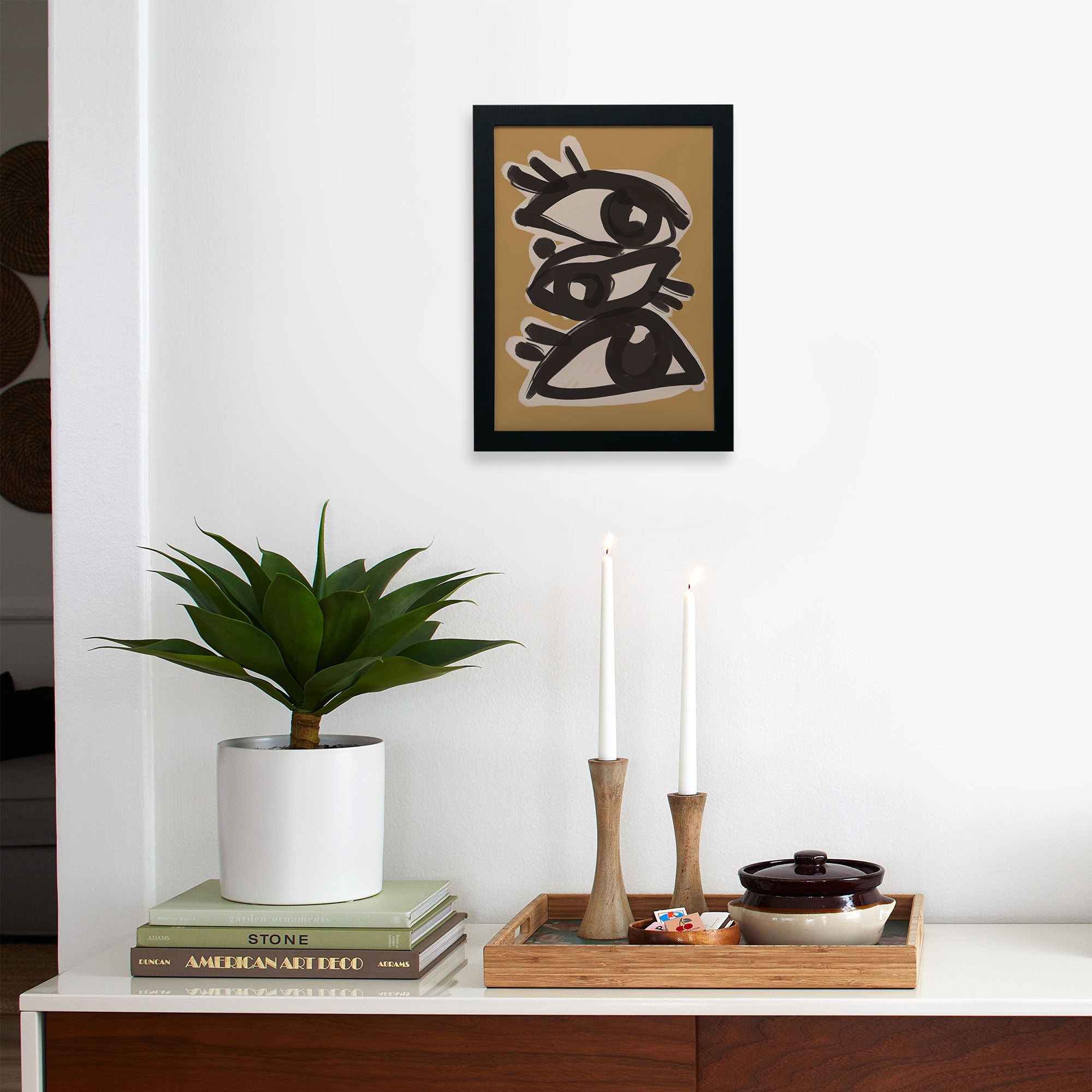 A modern, minimalist room displays a potted plant, books, and a tray with candles and a teapot. Above, Faire’s Abstract line art eyes—limited edition art on matte paper—depicts three outlined eyes on a tan background.