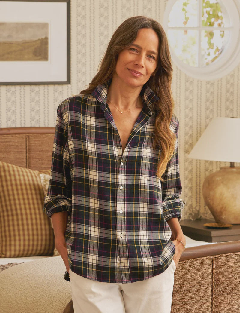 A woman with long brown hair wears the Frank & Eileen Eileen Button Up Shirt F25 and white pants, smiling indoors with hands in her pockets. The room features patterned wallpaper, a lamp, framed art, and neutral-toned decor.