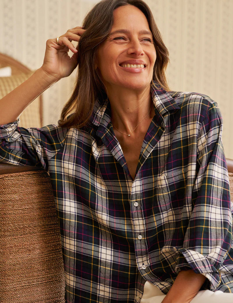 A woman with long brown hair smiles indoors, wearing the Frank & Eileen Eileen Button Up Shirt F25—a relaxed-fit plaid shirt in black, white, yellow, and red. She touches her hair and channels California style: confident, comfortable, and happy.