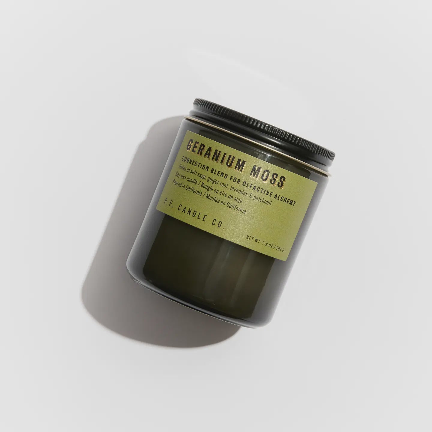 A dark green candle jar with a black lid labeled in bold, "Geranium Moss - 7.2 oz Alchemy Soy Candle" by Faire. Crafted from soy wax, it features scent notes and details on the label. Resting on a light surface casting a shadow, this candle captures sustainable patchouli elegance.