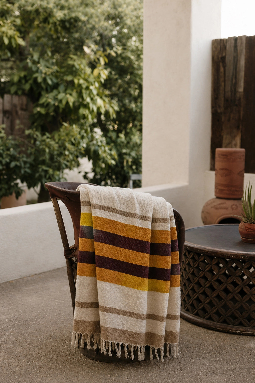 Handwoven striped Golden Horizon blanket by The Faire, draped on a patio chair near plants and a table.