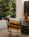Handwoven striped Golden Horizon blanket by The Faire, draped on a patio chair near plants and a table.