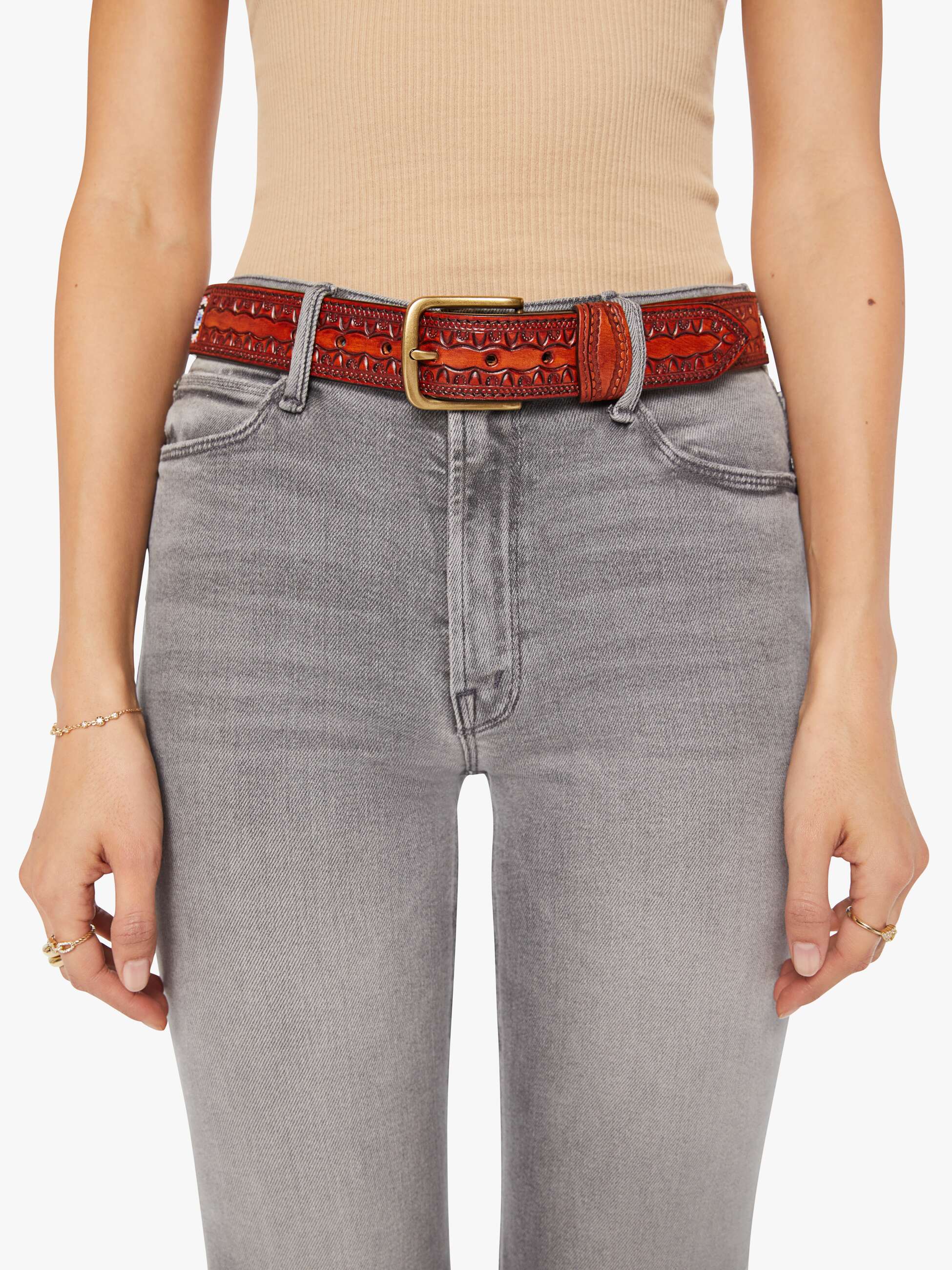 Wearing The Eye Opener beige sleeveless top by Mother with light gray high-waisted jeans and a vintage leather belt with brass buckle, hands relaxed at sides adorned with delicate rings and bracelets.