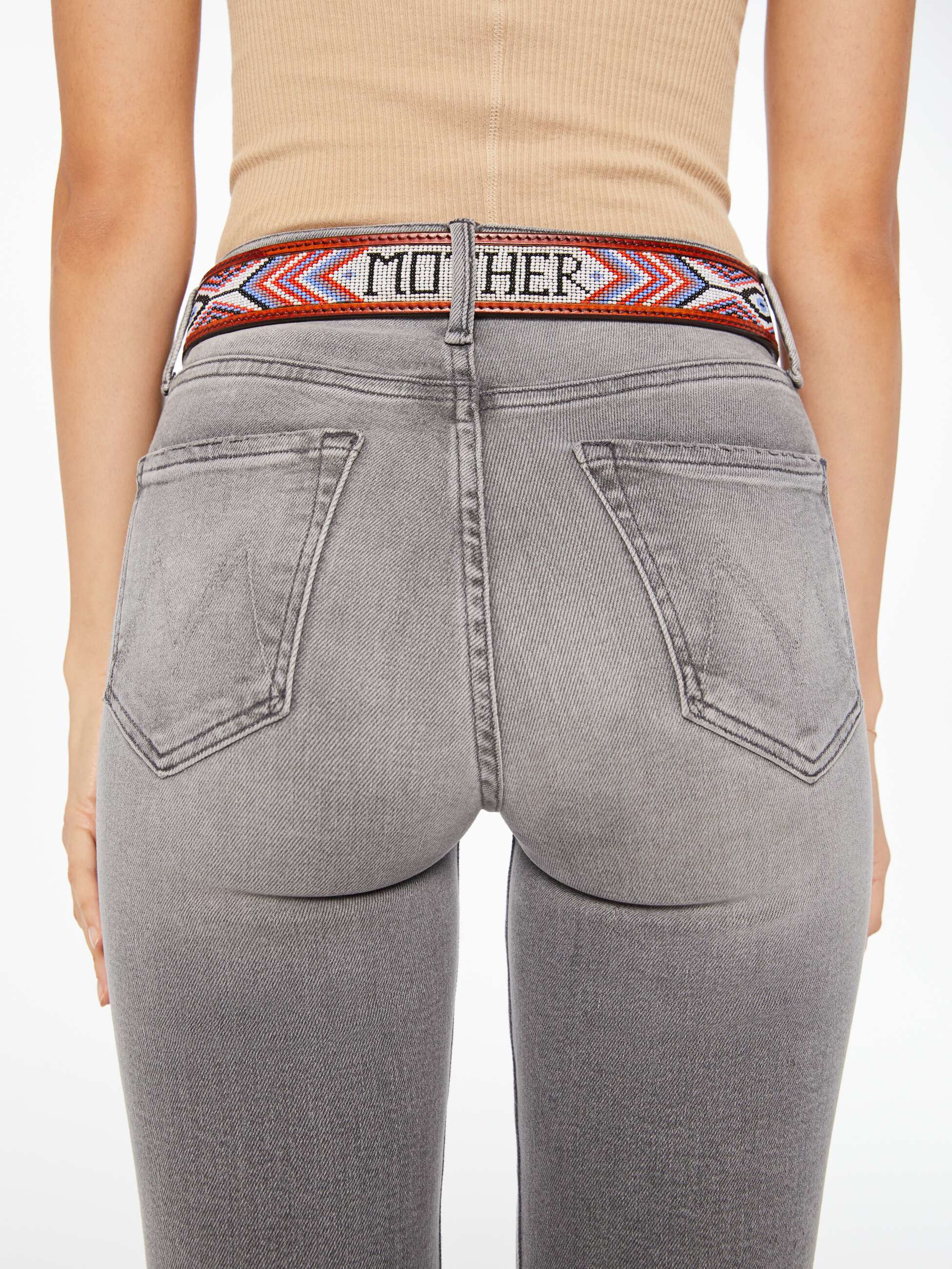 A person shown from the back wears The Eye Opener by Mother—a beige sleeveless top, gray jeans, and a colorful patterned beaded leather belt with "MOTHER" visible on the waistband.