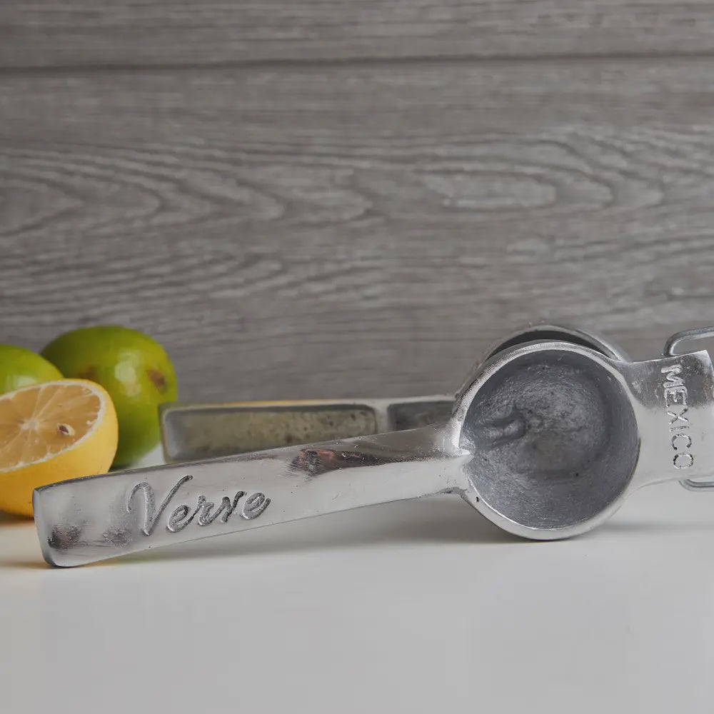 Faire&#39;s Aluminum Hand Juicer, expertly crafted in Mexico with &quot;Verve&quot; on the handle, rests on a gray surface. It&#39;s poised alongside a halved lemon and a whole lime, ready for juicing against the gray wooden backdrop.