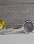 Faire's Aluminum Hand Juicer, expertly crafted in Mexico with "Verve" on the handle, rests on a gray surface. It's poised alongside a halved lemon and a whole lime, ready for juicing against the gray wooden backdrop.