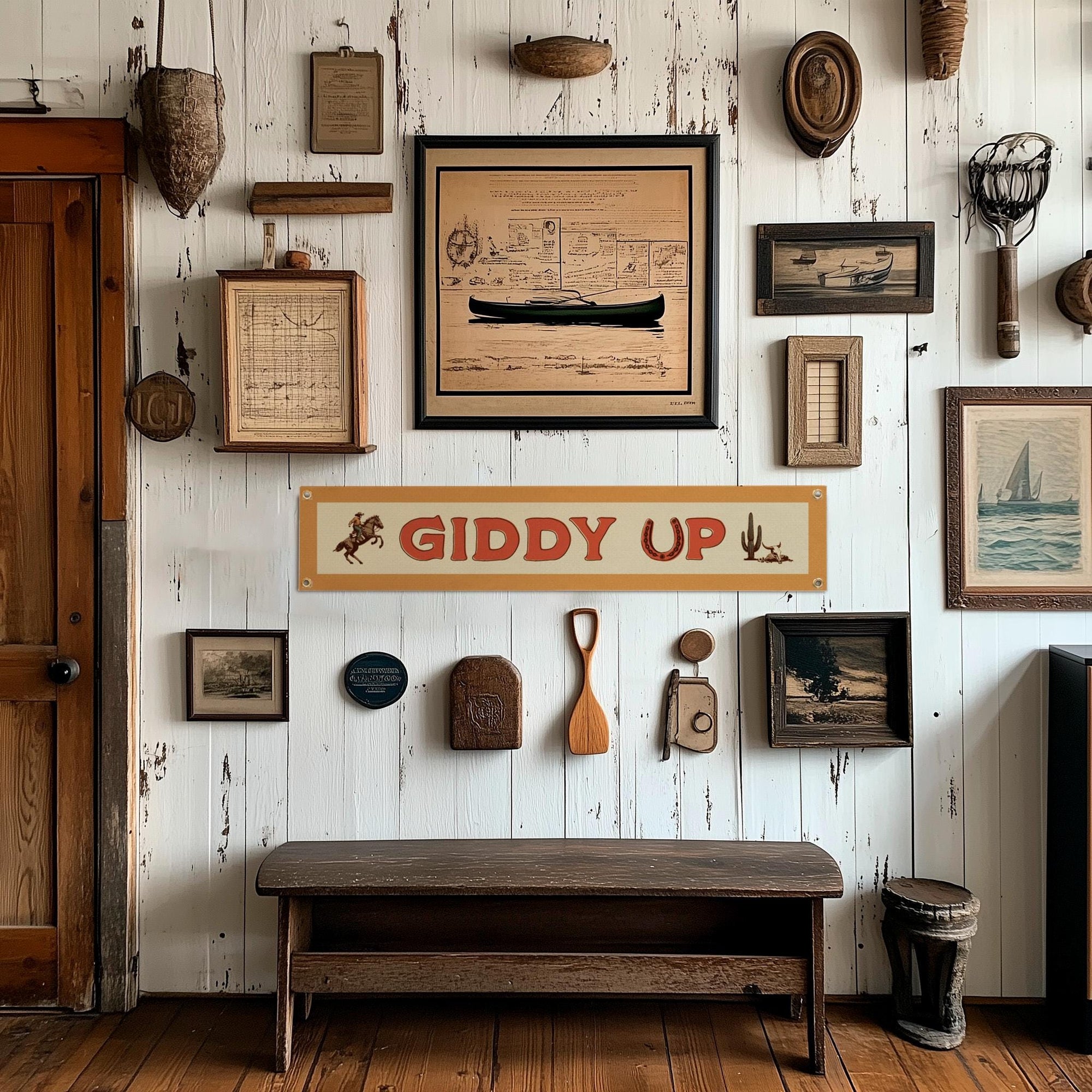 A rustic white wall displays vintage decor—frames, maps, paddles, fishing gear, and the Faire "Giddy Up" Felt Banner—above a wooden bench on polished floors. Sunlight enhances the space’s cozy, nostalgic charm.