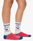 Two legs wear The Half-Calf Swim Club socks by Mother—white tube socks with blue and red stripes, pink toes, blue heels, and bold blue graphics of hands spelling “MOTHER” on one and “FUCKER” on the other.