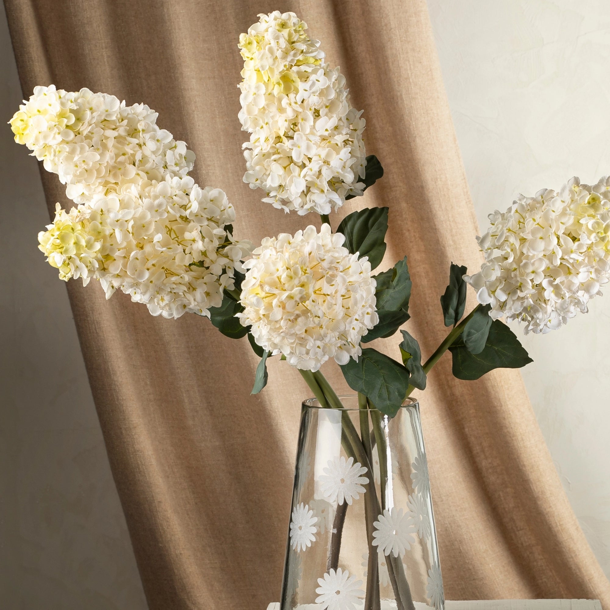 The Indaba Hydrangea Paniculata Stem features five lifelike faux stems arranged in a clear glass vase with white daisy patterns, displayed against a beige curtain and light-colored wall.